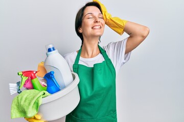 Young brunette woman with short hair wearing apron holding cleaning products smiling confident touching hair with hand up gesture, posing attractive and fashionable