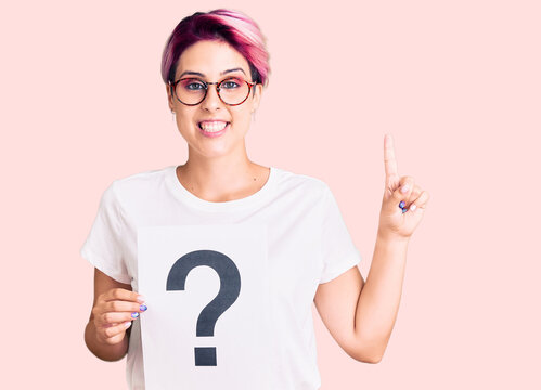 Young beautiful woman with pink hair holding question mark surprised with an idea or question pointing finger with happy face, number one