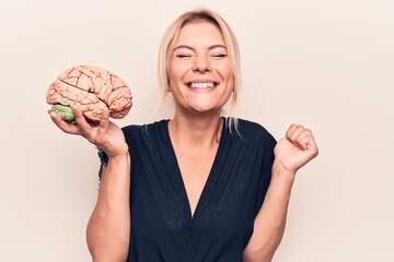 Young beautiful blonde woman asking for care memory holding brain over white background screaming proud, celebrating victory and success very excited with raised arm - obrazy, fototapety, plakaty