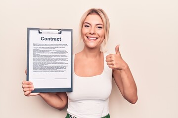 Beautiful blonde woman holding clipboard with contract document paper over white background smiling happy and positive, thumb up doing excellent and approval sign