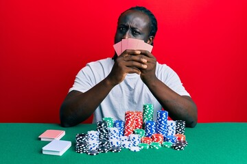 Handsome young black man playing gambling poker covering face with cards in shock face, looking skeptical and sarcastic, surprised with open mouth