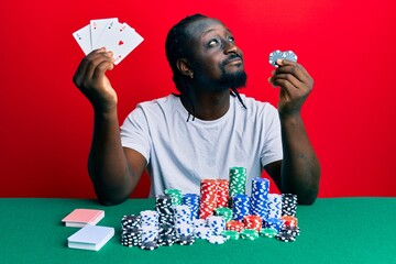 Handsome young black man sitting on the table with poker chips and cards smiling looking to the...