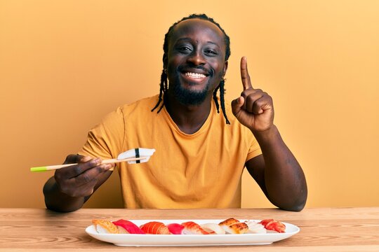 Handsome young black man eating sushi sitting on the table smiling with an idea or question pointing finger with happy face, number one