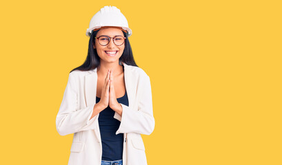Young beautiful latin girl wearing architect hardhat and glasses praying with hands together asking for forgiveness smiling confident.