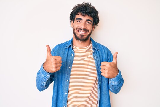 Handsome young man with curly hair and bear wearing casual denim jacket success sign doing positive gesture with hand, thumbs up smiling and happy. cheerful expression and winner gesture.