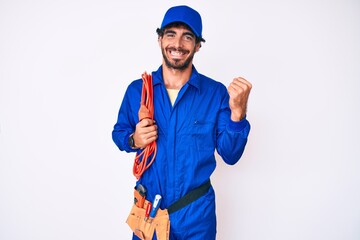 Handsome young man with curly hair and bear wearing builder jumpsuit uniform and electric cables...
