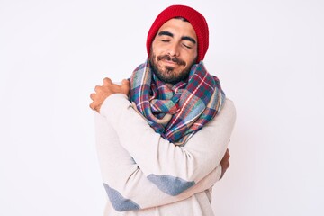 Fototapeta premium Handsome young man with curly hair and bear wearing a winter sweater, scarf and wool hat hugging oneself happy and positive, smiling confident. self love and self care