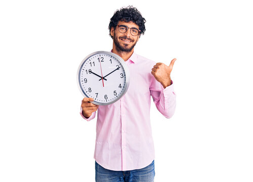 Handsome young man with curly hair and bear holding big clock pointing thumb up to the side smiling happy with open mouth