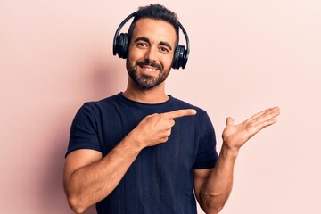 Young hispanic man listening to music using headphones amazed and smiling to the camera while presenting with hand and pointing with finger.