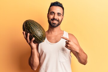 Young hispanic man holding melon smiling happy pointing with hand and finger