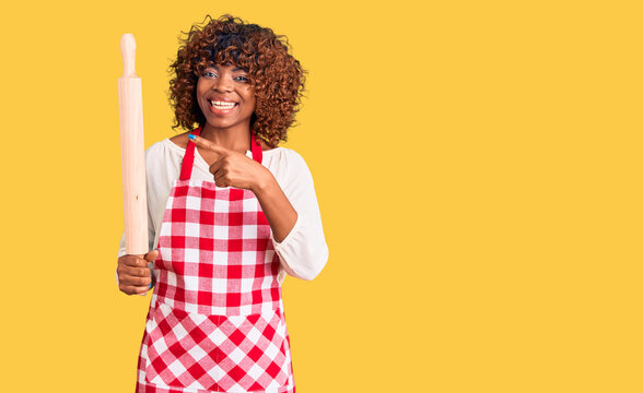 Young african american woman wearing apron holding rolling pin smiling happy pointing with hand and finger