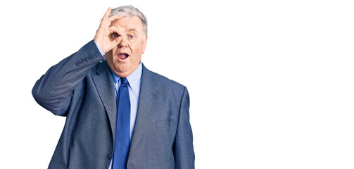 Senior grey-haired man wearing business jacket doing ok gesture shocked with surprised face, eye looking through fingers. unbelieving expression.