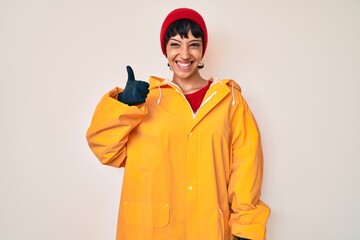 Beautiful brunettte woman wearing yellow raincoat smiling happy and positive, thumb up doing excellent and approval sign
