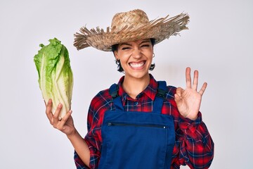 Beautiful brunettte woman wearing farmer clothes holding lettuce doing ok sign with fingers, smiling friendly gesturing excellent symbol