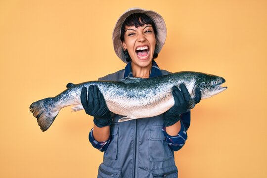 Beautiful brunettte fisher woman showing raw salmon celebrating crazy and amazed for success with open eyes screaming excited.