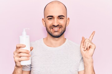 Young handsome bald man holding bottle of cream lotion smiling happy pointing with hand and finger to the side