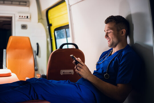 Young doctor sitting with his smartphone in the ambulance car at night.