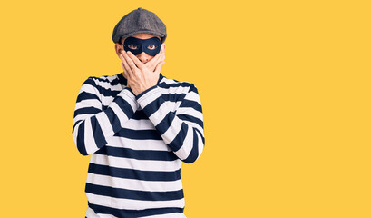 Young handsome man wearing burglar mask shocked covering mouth with hands for mistake. secret concept.