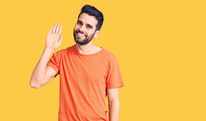 Young handsome man with beard wearing casual t-shirt waiving saying hello happy and smiling, friendly welcome gesture