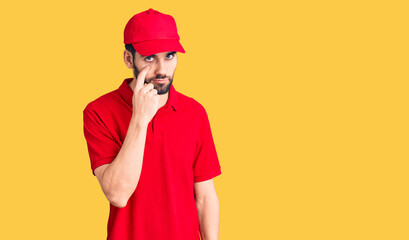 Young handsome man with beard wearing delivery uniform pointing to the eye watching you gesture, suspicious expression