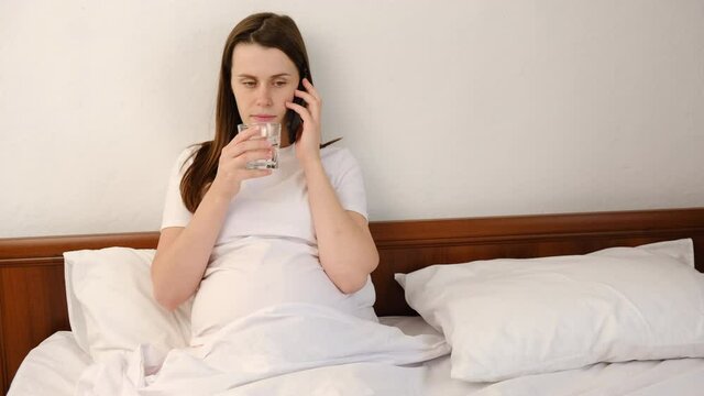 Young pregnant woman sitting on comfortable white bed use of mobile phone with glass and drink with water at home. Attractive future mom having pleasant conversation chatting by mobile with friend