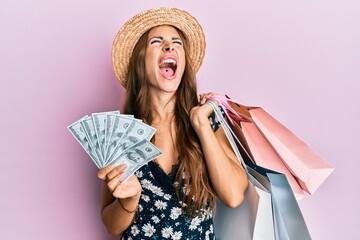 Young brunette woman holding shopping bags and dollars angry and mad screaming frustrated and furious, shouting with anger looking up.