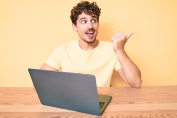 Young caucasian man with curly hair working at the office with laptop pointing thumb up to the side smiling happy with open mouth
