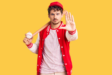 Young handsome man with curly hair playing baseball holding bat and ball with open hand doing stop sign with serious and confident expression, defense gesture