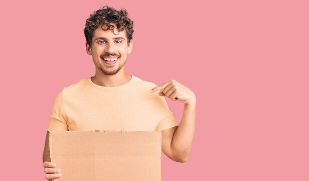 Young handsome man with curly hair holding blank cardboard empty banner pointing finger to one self smiling happy and proud