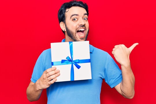 Young hispanic man holding gift pointing thumb up to the side smiling happy with open mouth