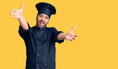 Young hispanic man wearing cooker uniform looking at the camera smiling with open arms for hug....