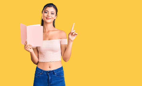 Young beautiful woman holding book surprised with an idea or question pointing finger with happy face, number one