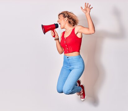 Young beautiful caucasian woman screaming using megaphone. Jumping over isolated white background