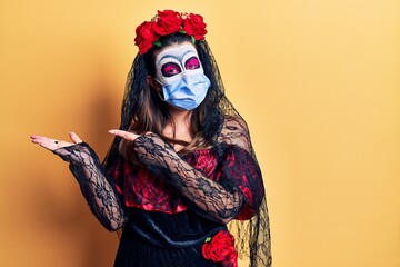 Young woman wearing day of the dead costume wearing medical mask amazed and smiling to the camera while presenting with hand and pointing with finger.