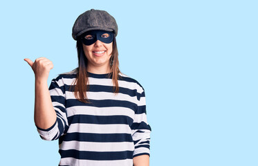 Young beautiful brunette woman wearing burglar mask smiling with happy face looking and pointing to the side with thumb up.