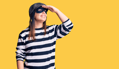 Young beautiful brunette woman wearing burglar mask very happy and smiling looking far away with hand over head. searching concept.