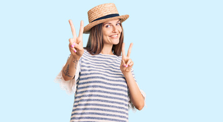 Beautiful caucasian woman wearing summer hat smiling looking to the camera showing fingers doing victory sign. number two.