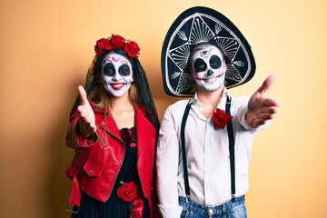 Couple wearing day of the dead costume over yellow smiling friendly offering handshake as greeting and welcoming. successful business.