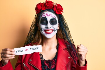 Woman wearing day of the dead costume holding trick or treat paper screaming proud, celebrating victory and success very excited with raised arm