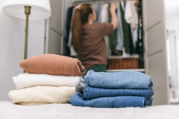 A woman is cleaning out her wardrobe, standing by clothes placed on hangers and in drawers. In the foreground are stack of sweaters and jeans