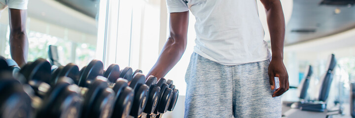 African American man picking up a dumbbell from the rack and looking outside at gym. Male weight training person holding sport equipment with serous look in fitness center