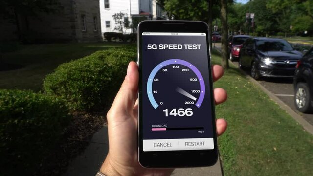 A man walks down the street in a small town while testing his 5G download bandwidth speed on a cellular phone. Fictional screen created in After Effects.  	