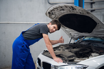A mechanic stands by the open hood of a car and shows a thumb