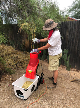 Adult male wearing thick protective gloves feeds garden sticks and small branches into an electric wood chipper.