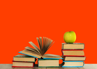 Open textbook, pile of books in colorful covers and green apple on wooden table with red orange background. Distance home education. Back to school, quarantine concept of stay home.