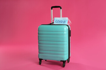 Stylish turquoise suitcase and protective masks with inscription COVID-19 on pink background. Travelling during coronavirus pandemic