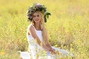 Young woman wearing wreath made of beautiful flowers in field on sunny day