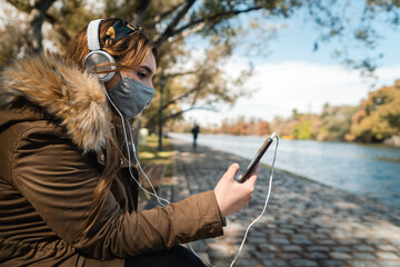 A caucasian woman wearing face mask and headphones relaxing and using her smartphone at park. Copy space