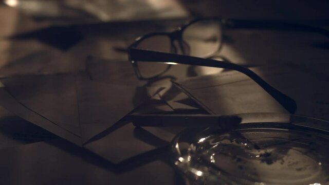 Cigarette On The Ashtray And Eye Glasses Lies On The Scattered Documents With A Bright Sunlight And Shadow Of Curtains Swaying On The Wind. - close up shot