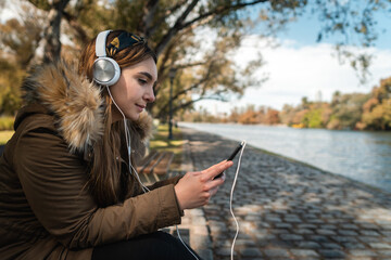 A beautiful woman wearing headphones and using her smartphone in front of a lake at park. Copy space